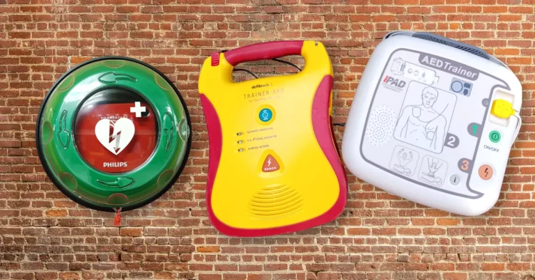Trzy różne modele defibrylatorów szkoleniowych AED (Philips w zielonej szafce ściennej, żółty Defibtech i biały iPAD Trainer) zawieszone na tle ceglanej ściany.