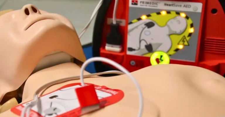 Zbliżenie na klatkę piersiową fantoma szkoleniowego z przyklejoną elektrodą defibrylatora. W tle widoczny podświetlony panel szkoleniowego urządzenia AED Primedic HeartSave.